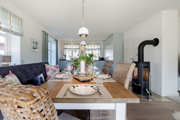 Un coin repas avec une table en bois dressée pour deux personnes, un poêle à bois noir et un canapé avec des coussins.