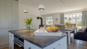 A modern kitchen with white cabinets, a central island with a bowl of lemons, pendant lights, and a view of the living area with a sofa and dining table.