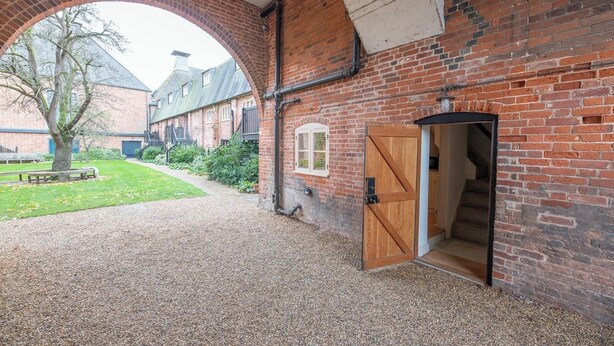 A brick building with a large arched entrance, a wooden door, and a gravel courtyard. - Kiln House, Snape Maltings (Snape)