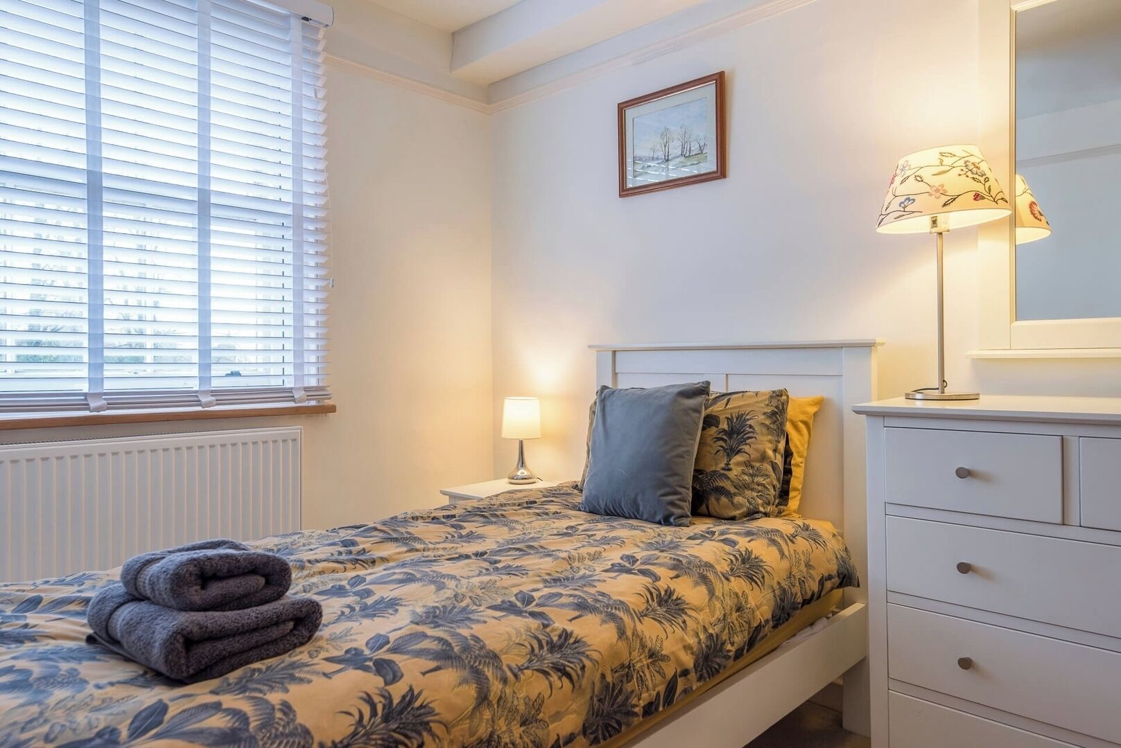 A neatly made bed with a floral bedspread, a bedside lamp, and a folded towel on the floor.
