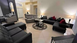 A spacious living room with a sectional sofa, a coffee table, and a TV. There is a door leading to another room and a lamp on a side table.