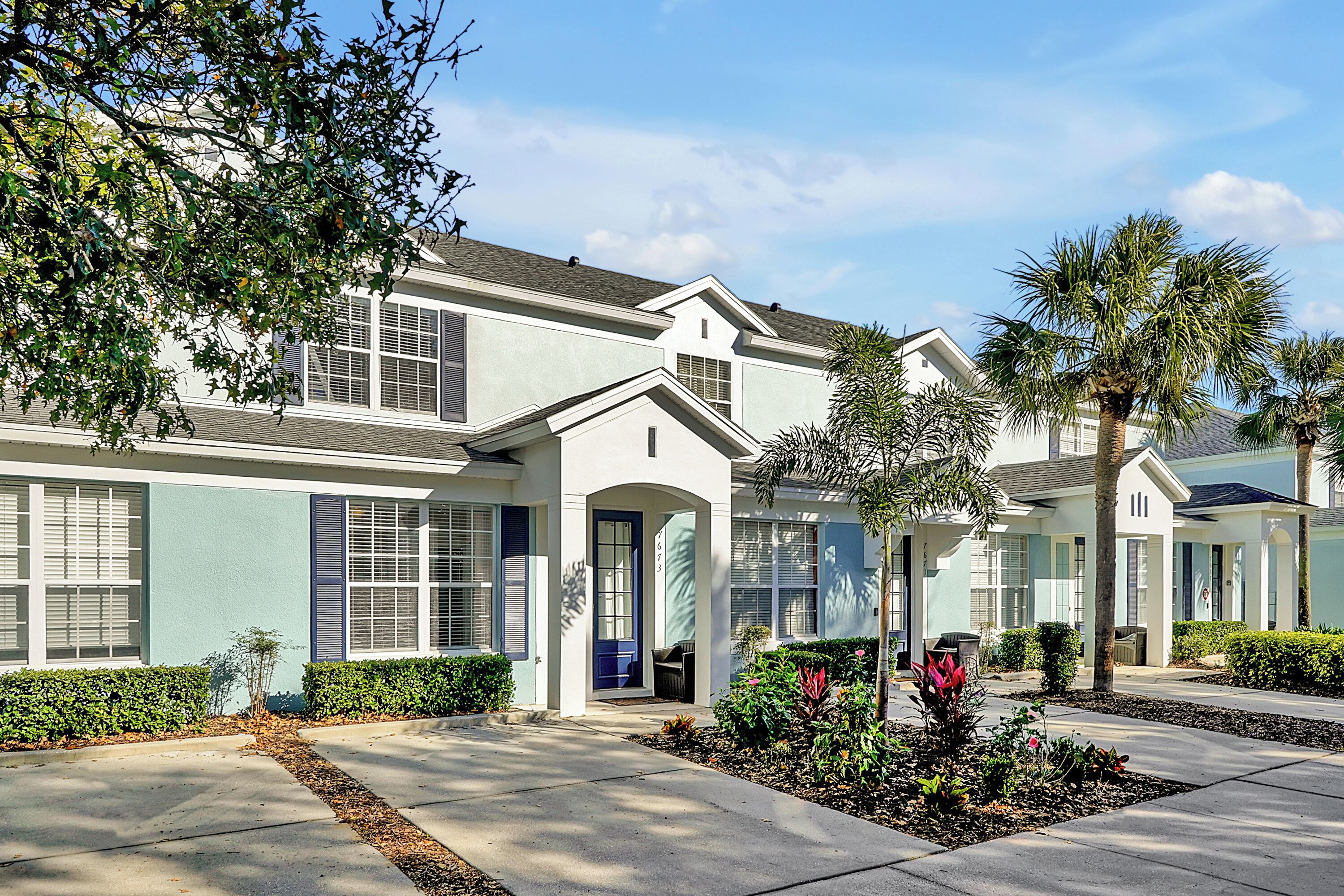 A modern two-story house with a light blue exterior, a front porch, and a landscaped garden with palm trees and shrubs.