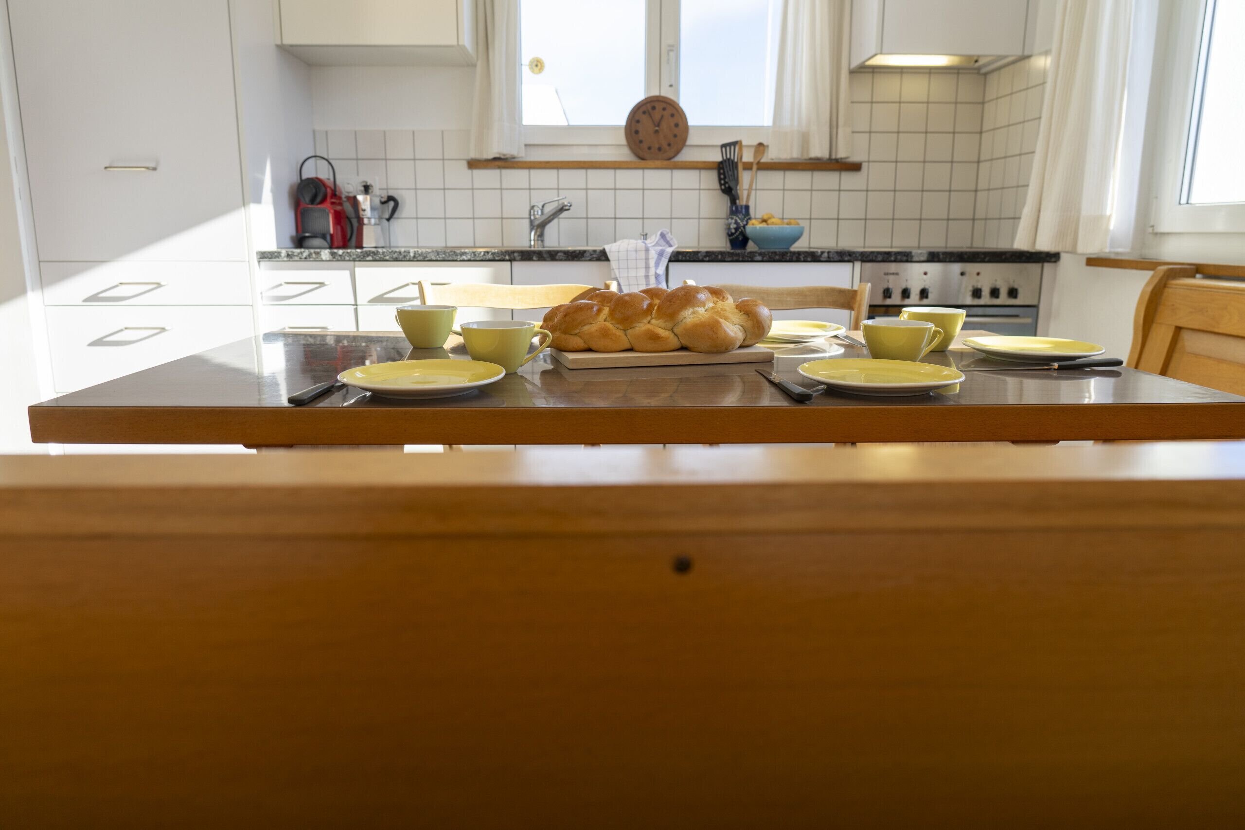A kitchen with a wooden table set for two, featuring a loaf of bread, yellow cups, and plates.