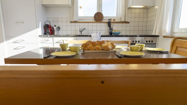 Una cocina con una mesa de madera dispuesta para dos personas, que incluye una hogaza de pan, tazas amarillas y platos.