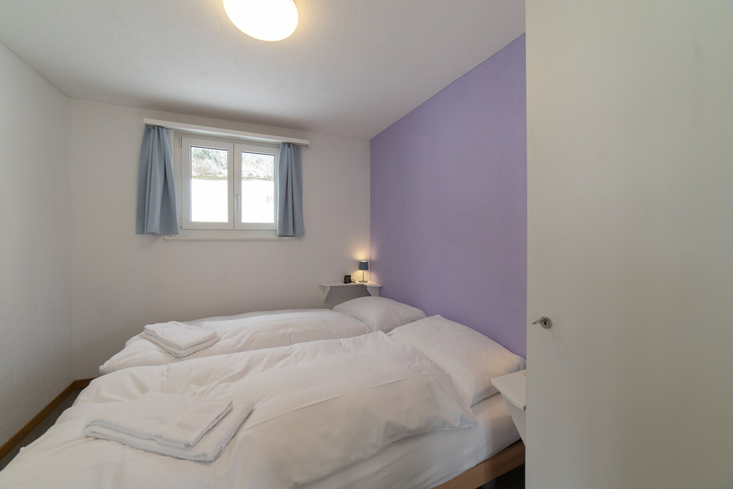 A bedroom with two beds, a window with curtains, a bedside table with a lamp, and a door.