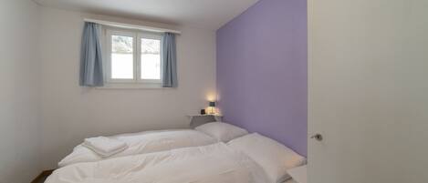 A bedroom with two beds, a window with curtains, a bedside table with a lamp, and a door.