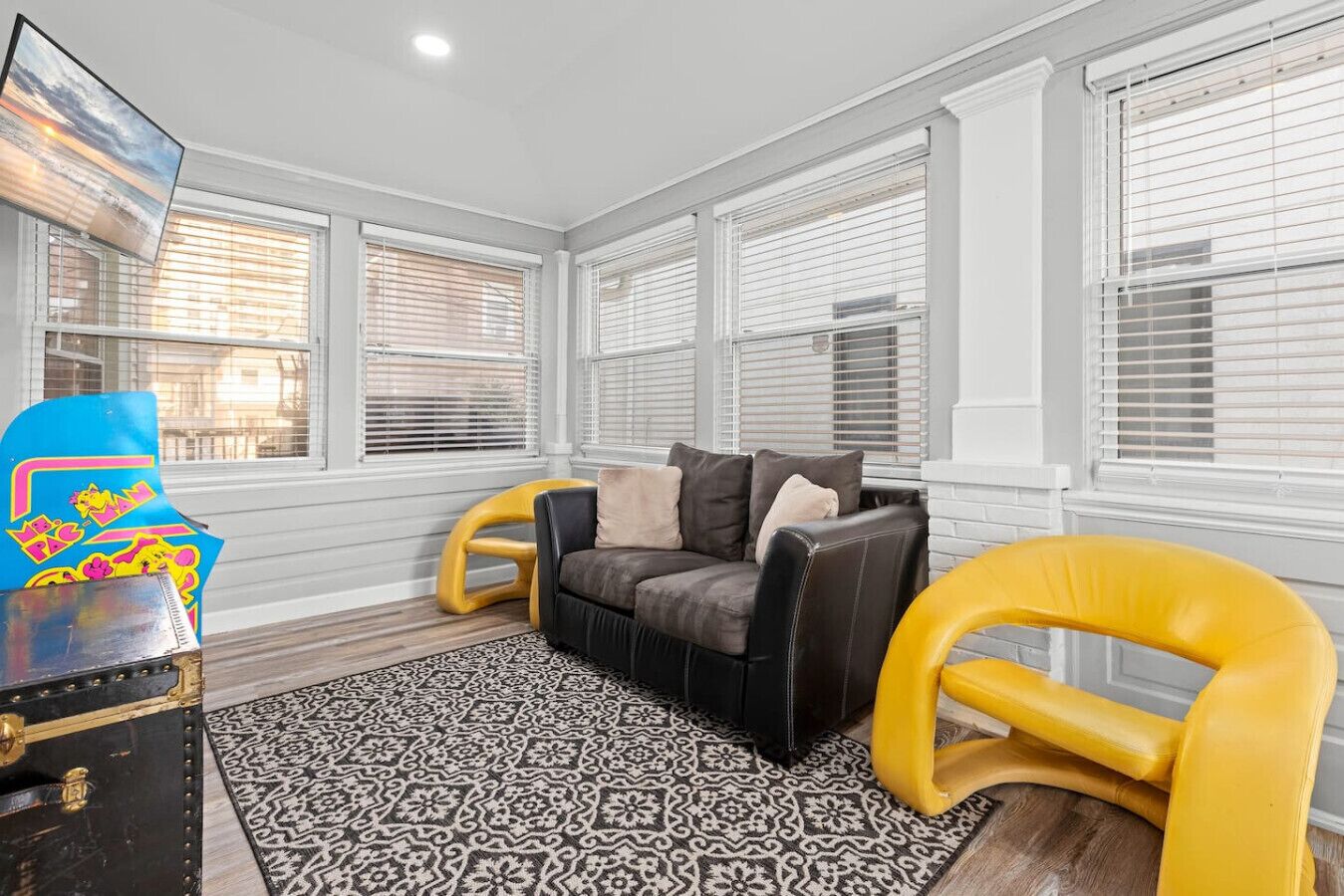 A living room with a black leather sofa, yellow armchairs, a patterned rug, and a large TV on the wall.
