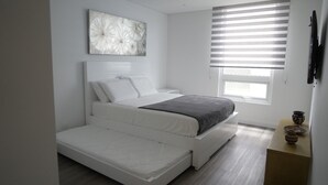 A modern bedroom with a bed, a window with blinds, and a small table with decorative items. - Modern new 3 bedroom apartment 5min walk to beachl/shops/restaurants (San Andrés)