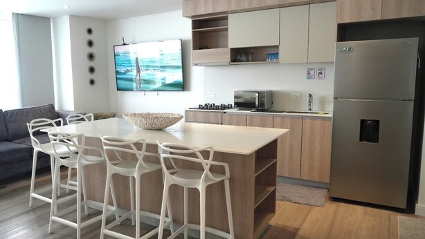 A modern kitchen with a central island, bar stools, a stainless steel refrigerator, and a flat-screen TV mounted on the wall. - Modern new 3 bedroom apartment 5min walk to beachl/shops/restaurants (San Andrés)