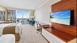 A hotel room with a bed, a desk, a chair, a TV on the wall, and a view of a city and water. - LUXURIOUS APARTMENT FRONT OF THE BEACH IN SUNNY ISLE (Sunny Isles Beach)