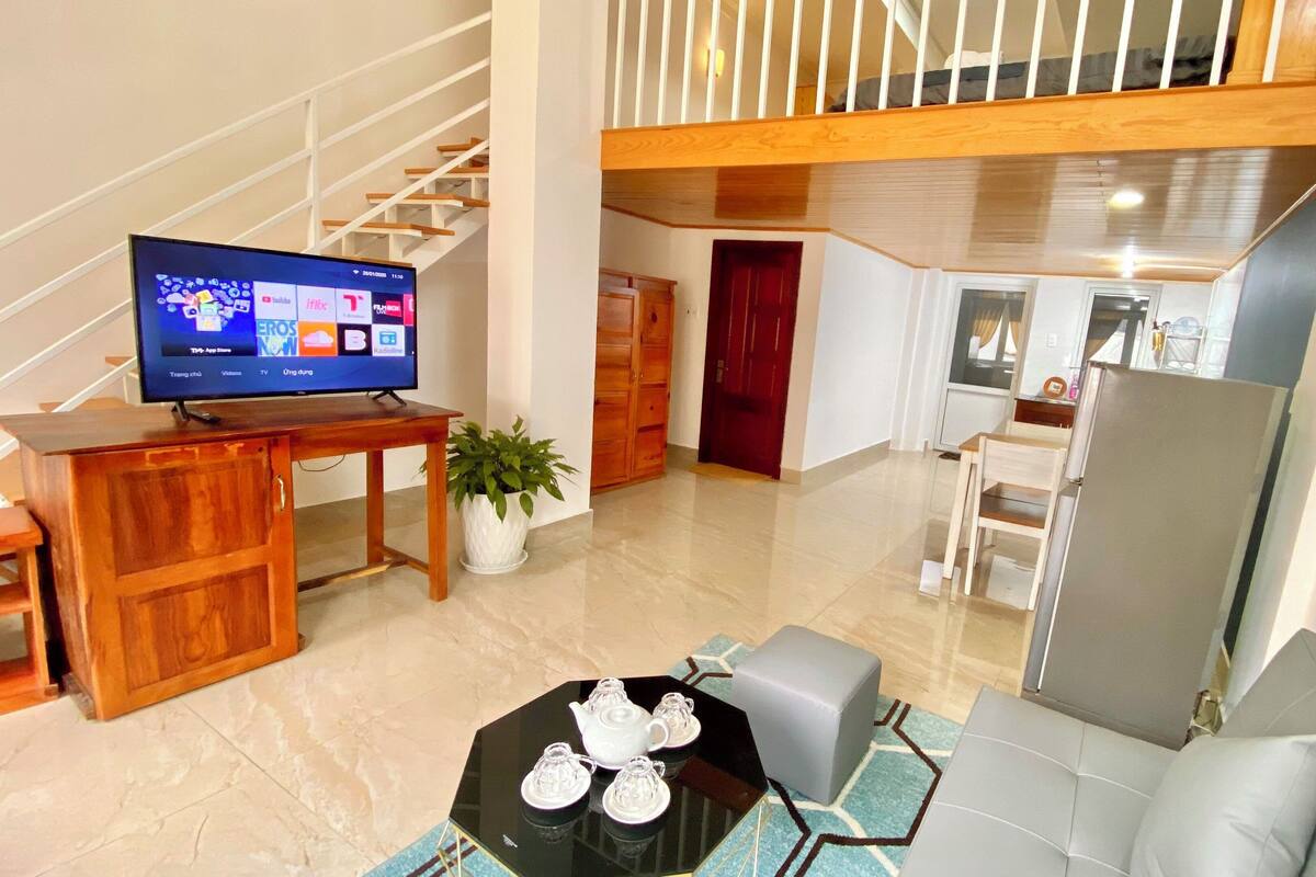 A modern living room with a TV, a dining area, a staircase, and a kitchen.