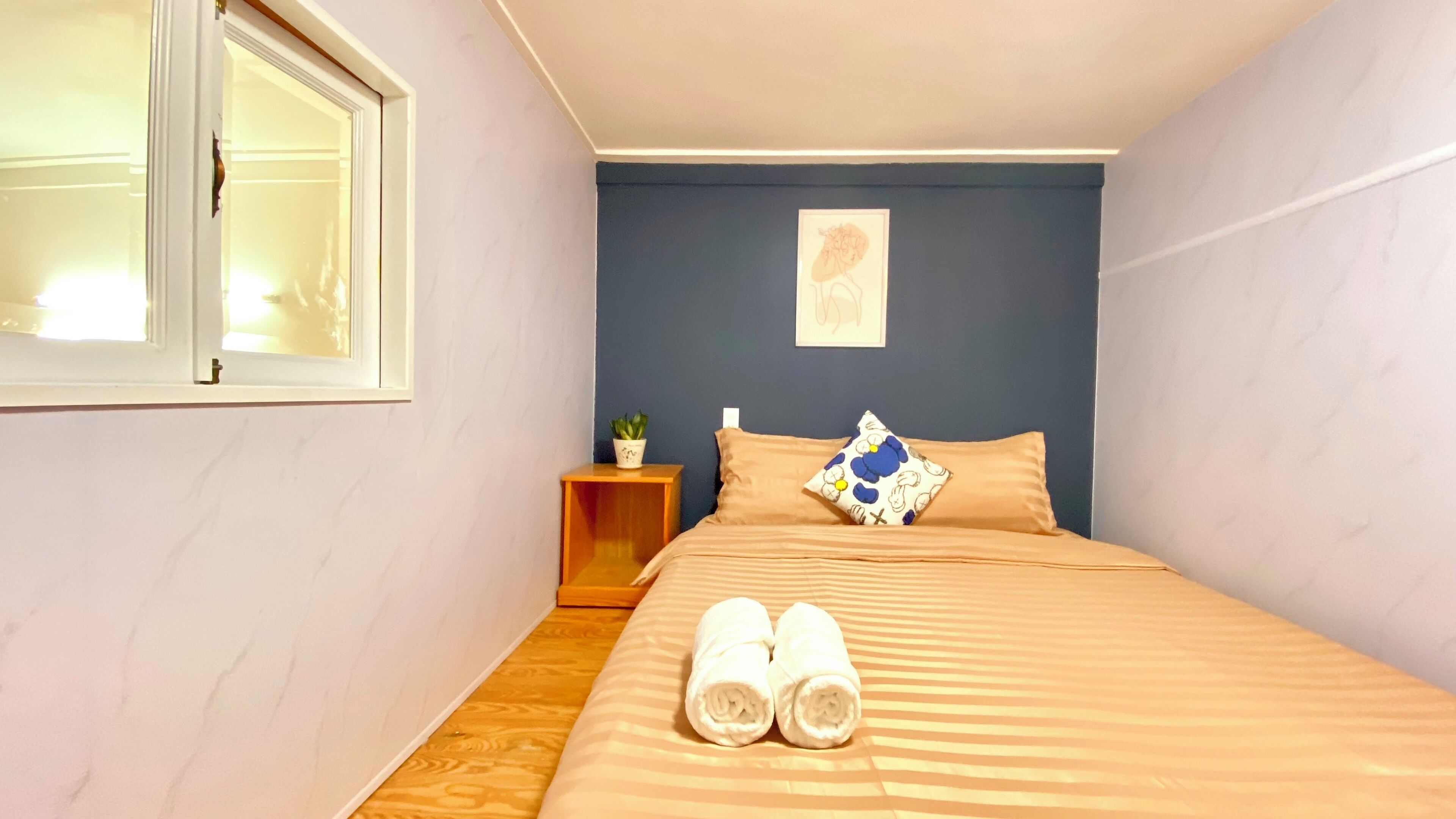 Une chambre à coucher avec un lit, des oreillers, une table de chevet, une plante en pot et une fenêtre.