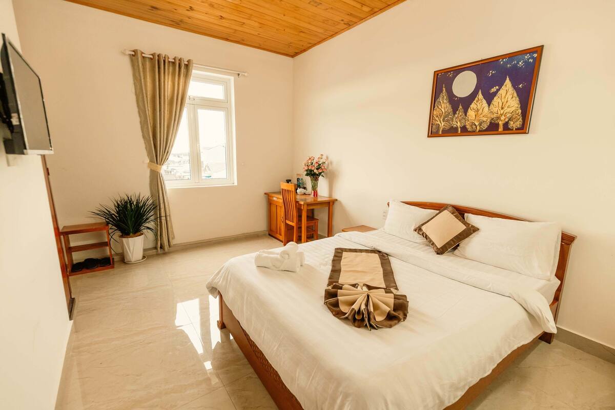 A neatly made bed with white linens and a decorative towel, a wooden headboard, a night sky painting, a potted plant, and a desk with a chair.
