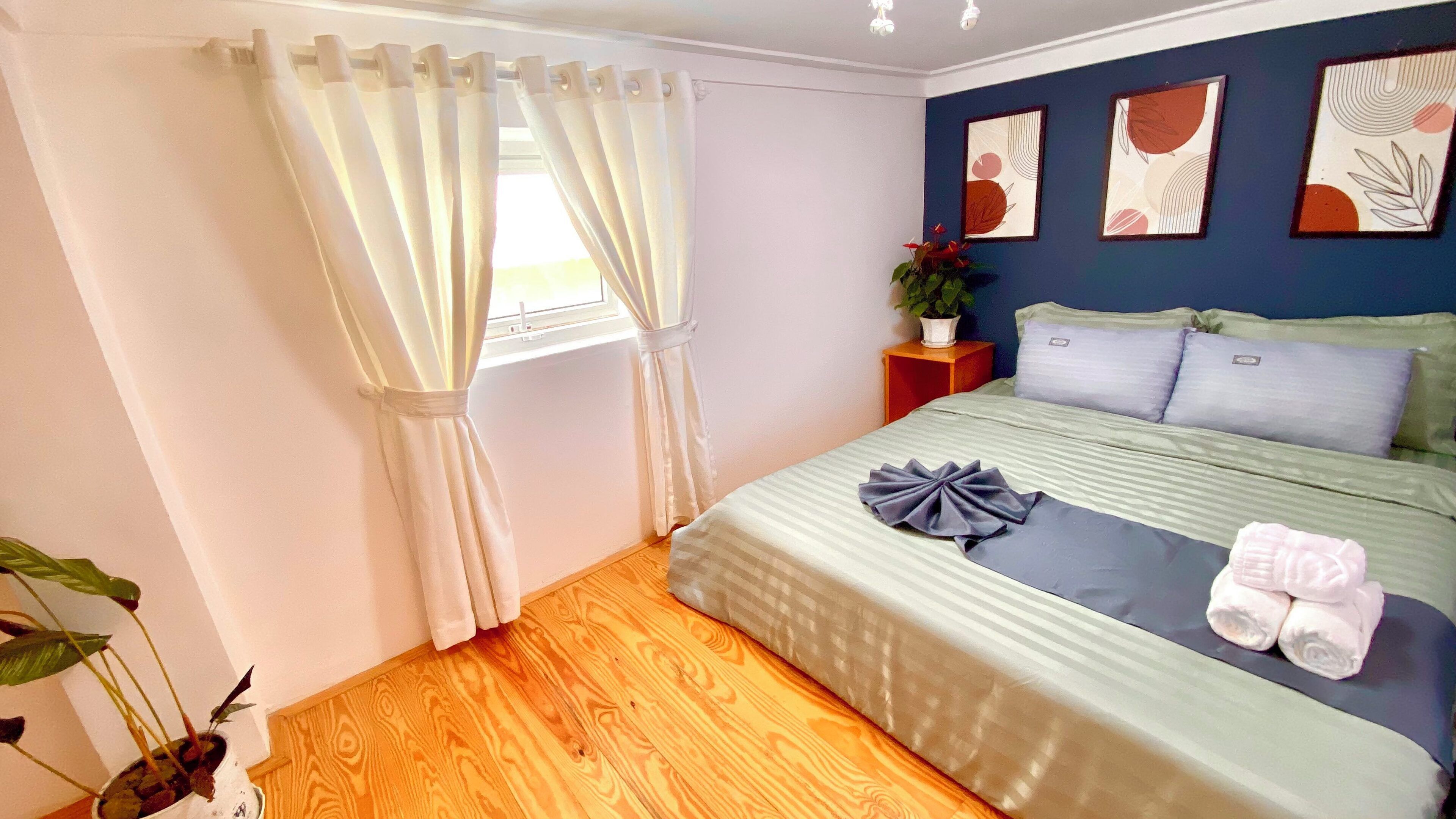 Une chambre à coucher avec un lit, des rideaux, une table de chevet, une plante en pot et des œuvres d’art mural.