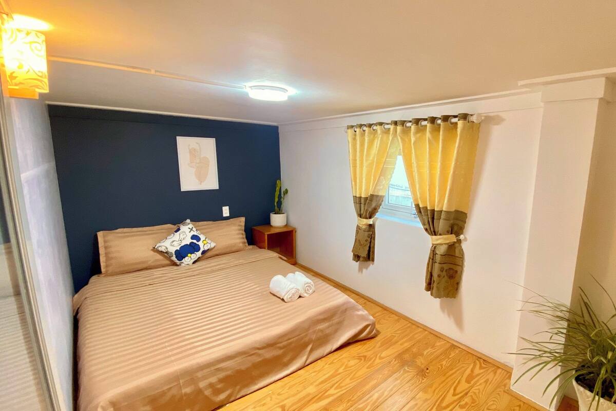 A bedroom with a bed, pillows, a nightstand, a potted plant, and curtains.