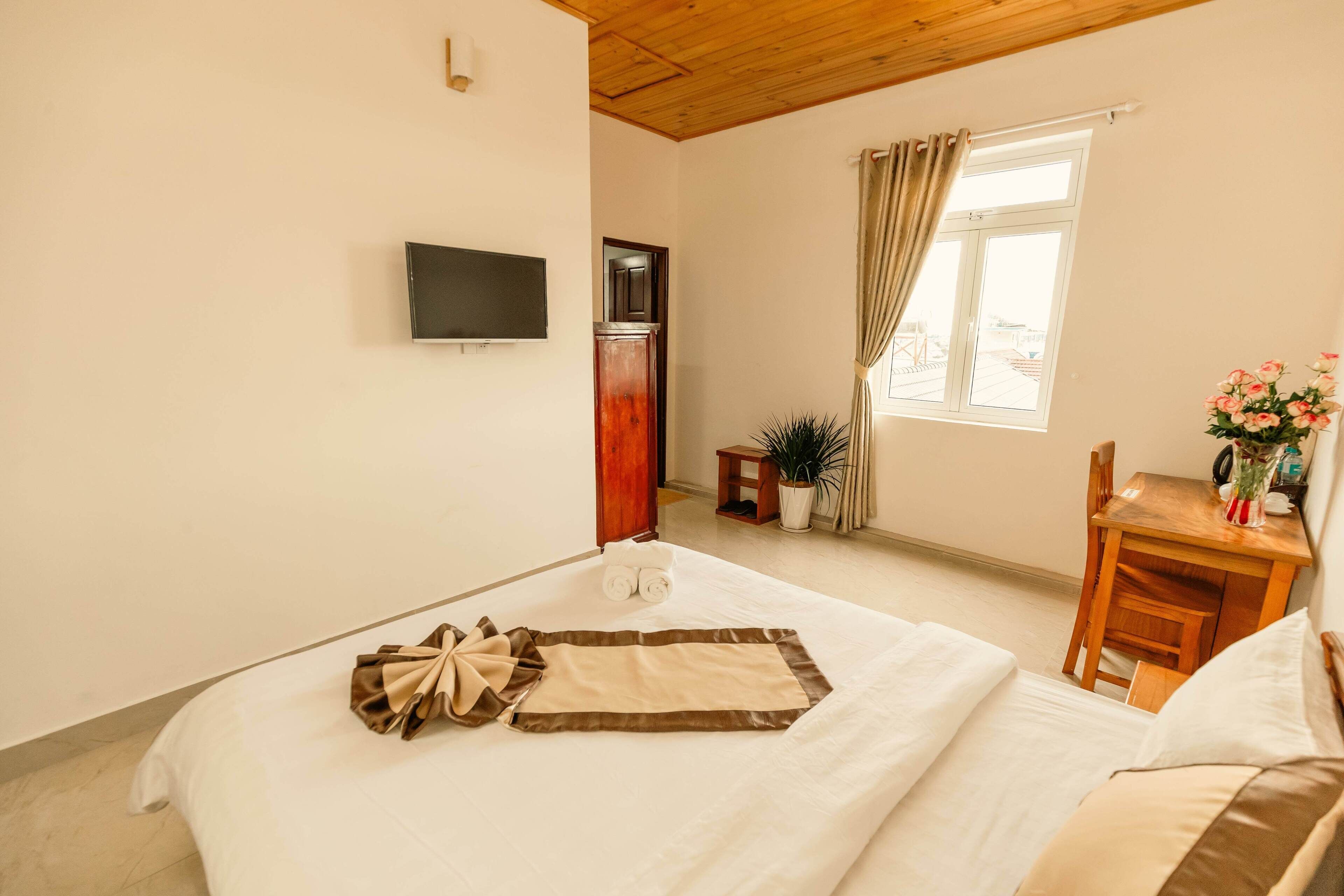 A hotel room with a bed, a TV mounted on the wall, a desk with a chair, a window with curtains, and a potted plant.