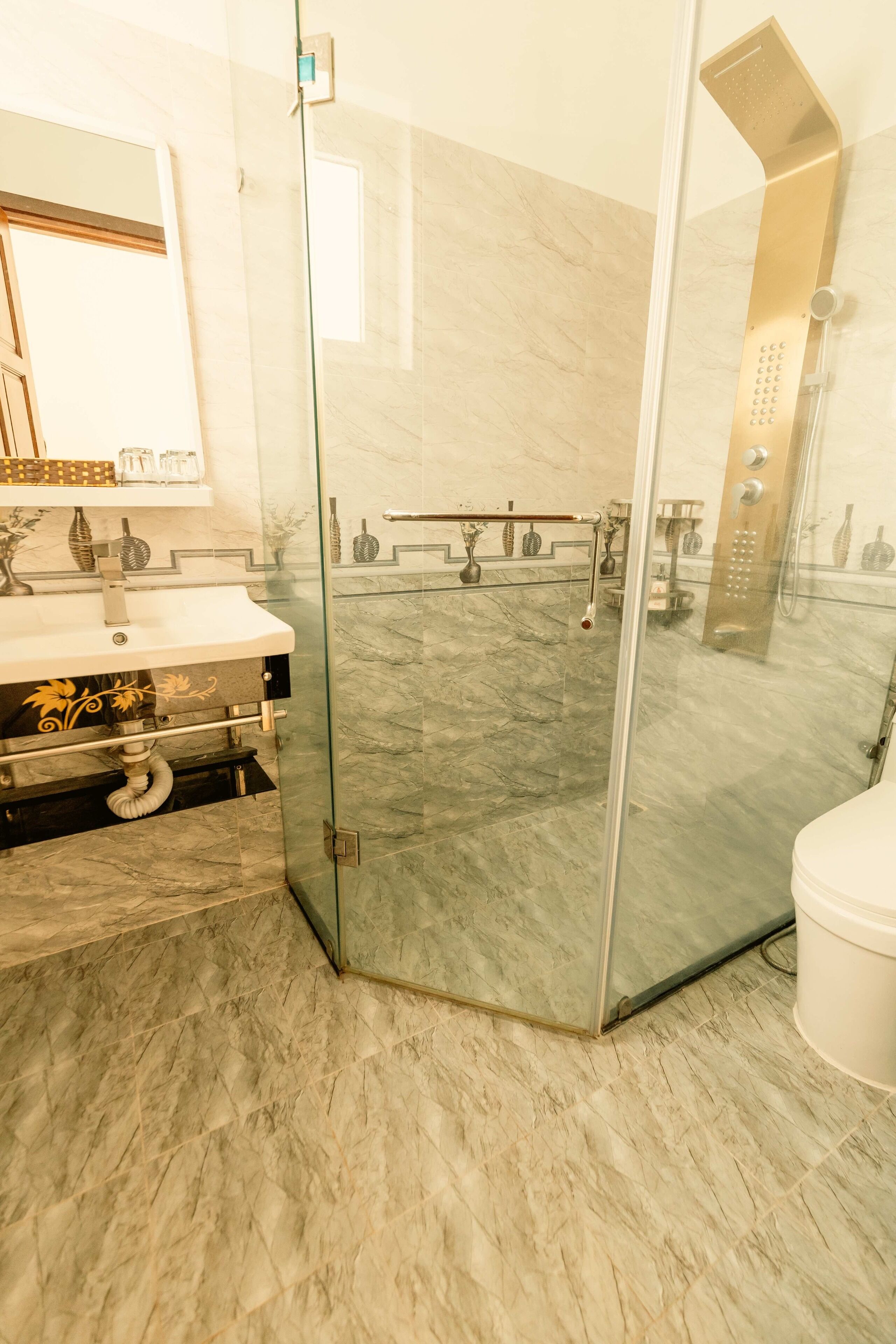 A modern bathroom with a glass-enclosed shower, a marble floor, and a white sink with a gold decorative panel.