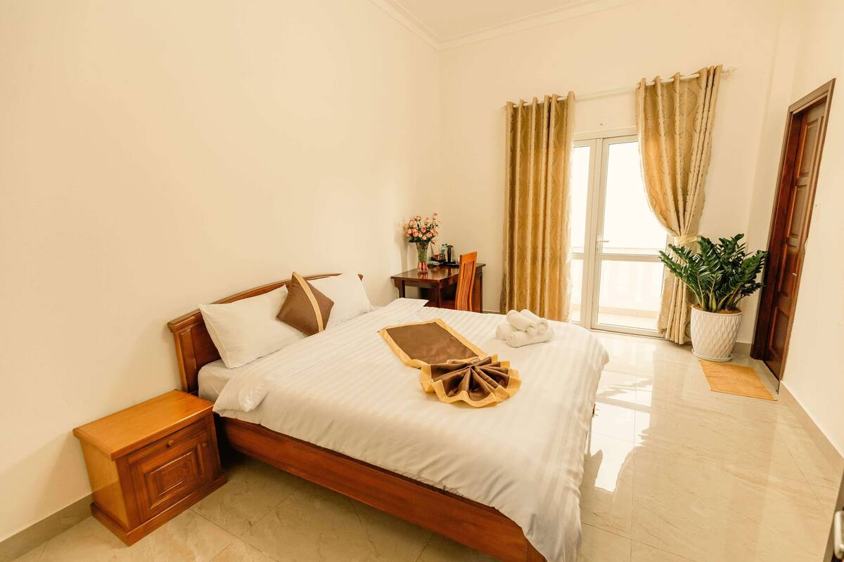 A neatly made bed with a white bedspread, a wooden nightstand, a small table with a vase of flowers, and a potted plant near the window.