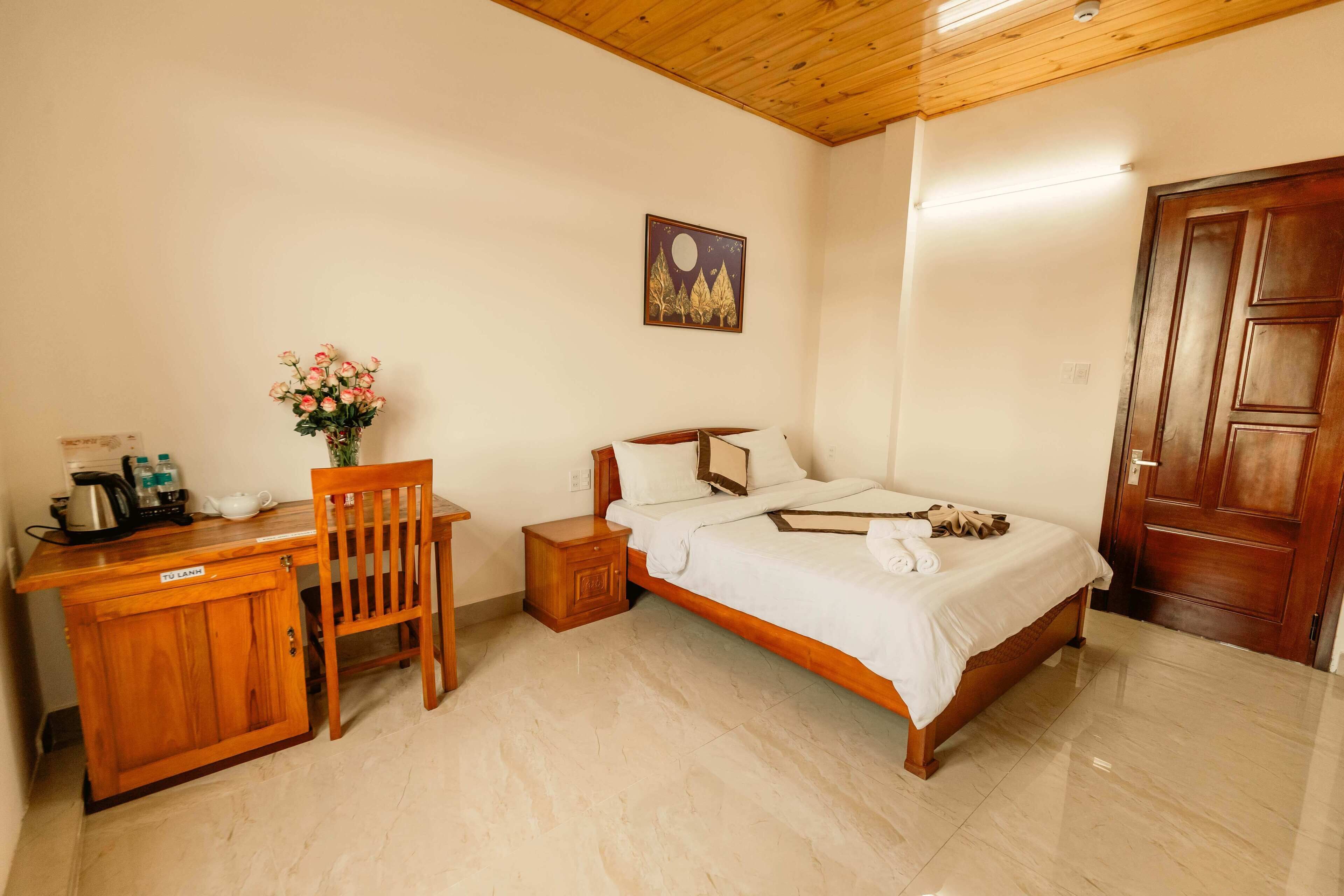 A bedroom with a wooden bed, a desk, a chair, a nightstand, and a wall-mounted painting.