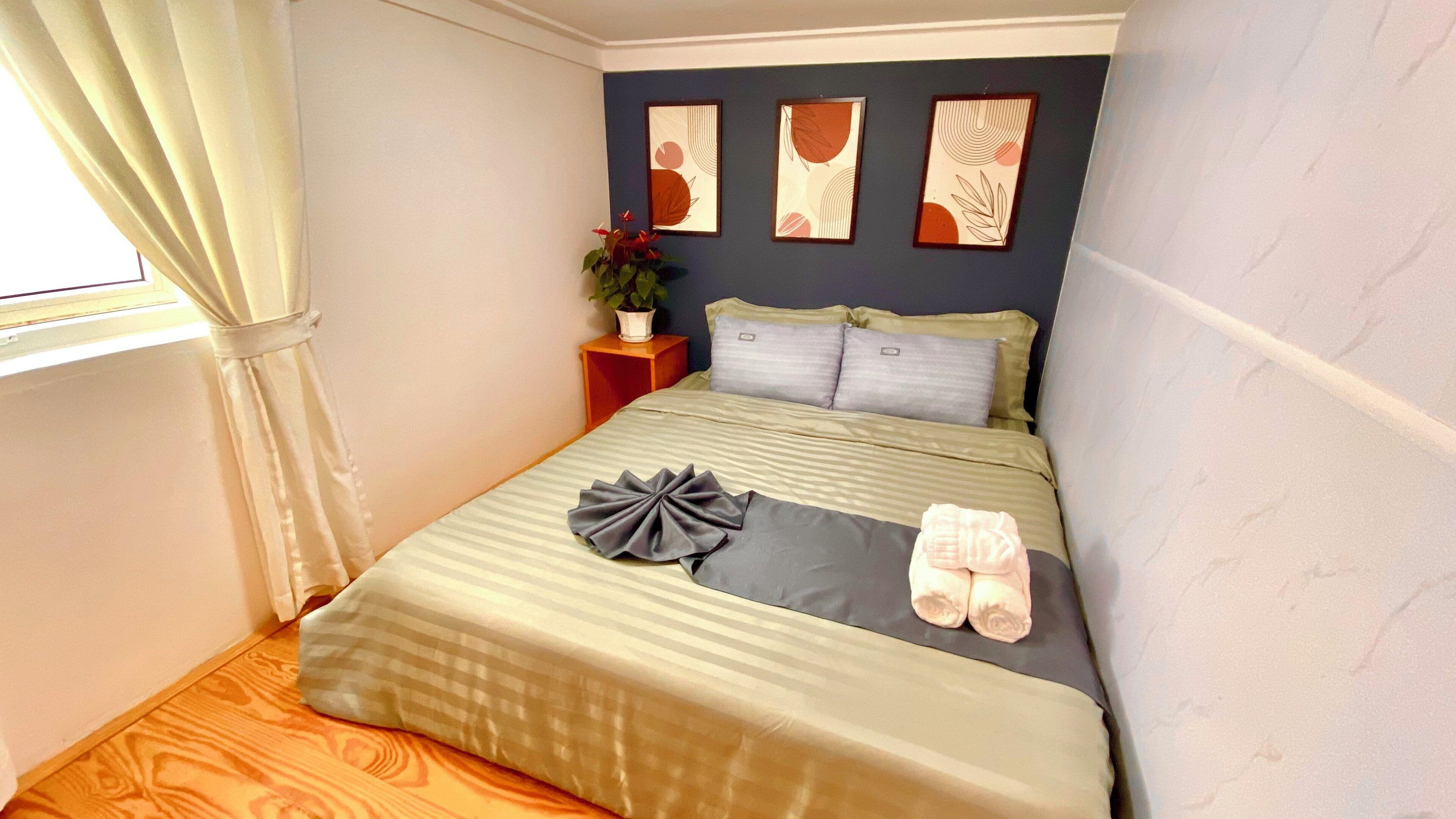 A neatly made bed with a striped bedspread, two pillows, and a decorative bow. A potted plant is on a nightstand beside the bed.