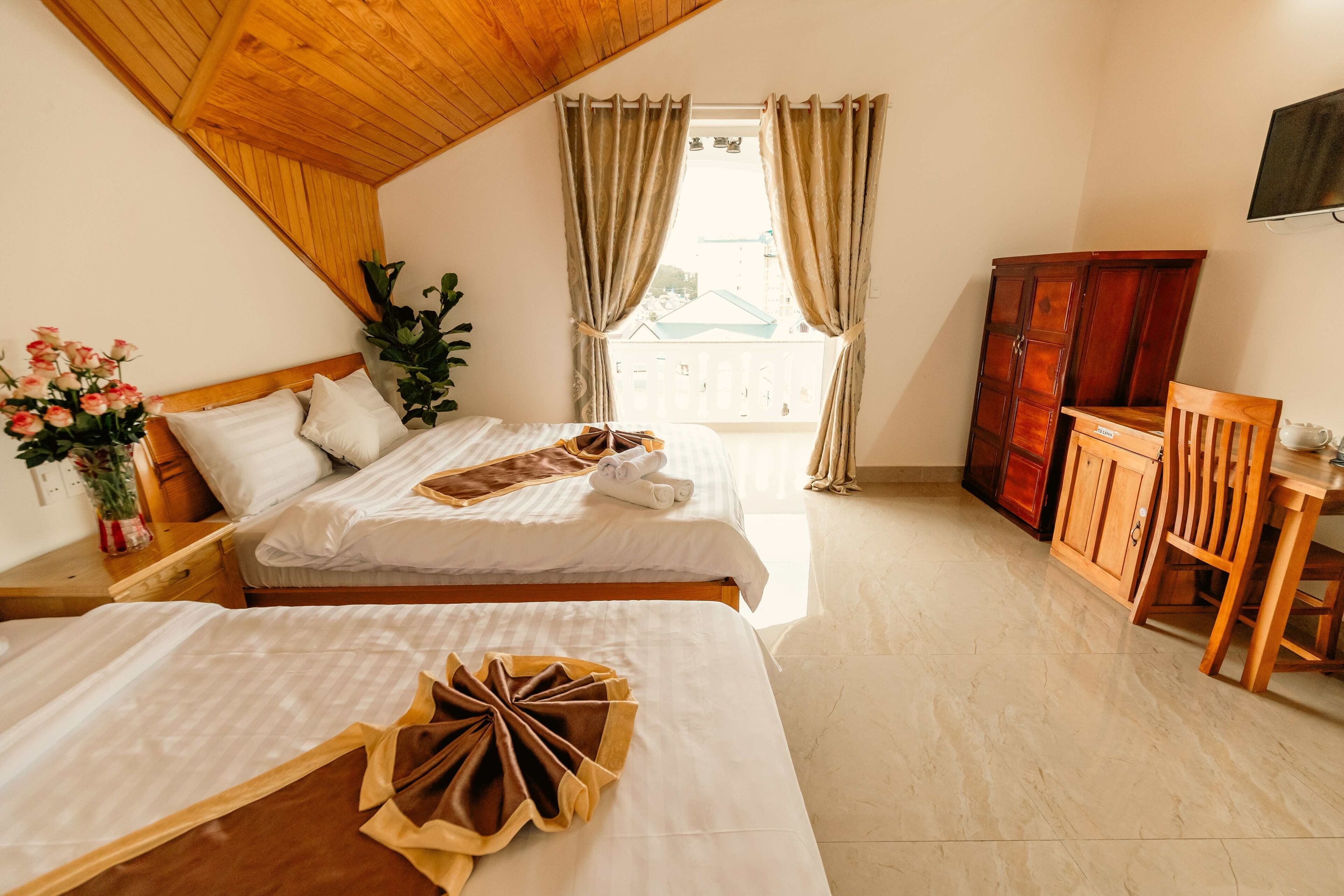 A hotel room with two beds, a wooden ceiling, a large window with curtains, a wooden cabinet, and a TV mounted on the wall.