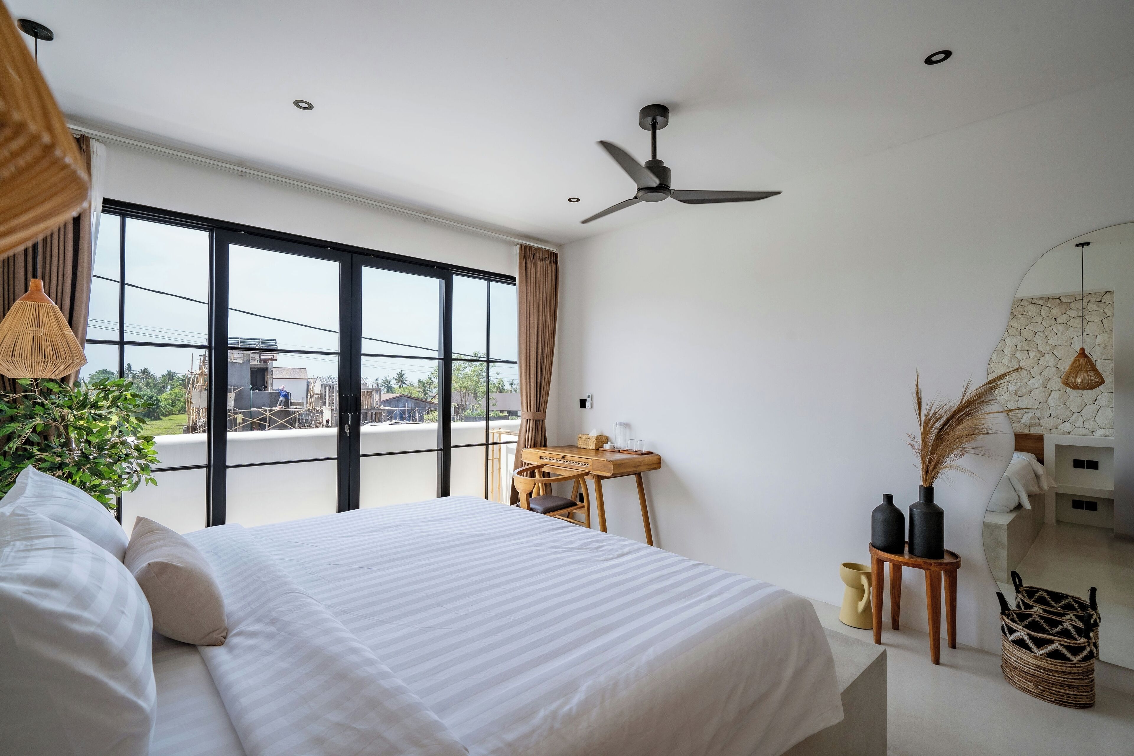 A modern bedroom with a large bed, a ceiling fan, a desk, and a view through large windows.