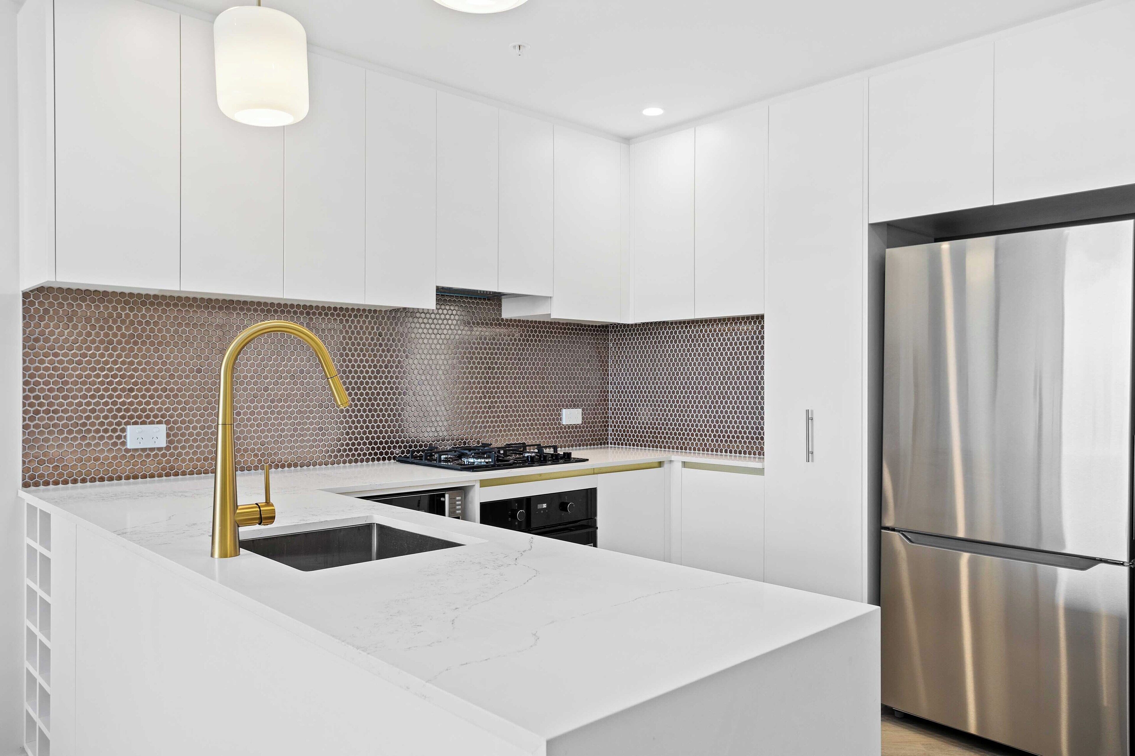 A modern kitchen with white cabinetry, a central island with a gold faucet, a stainless steel refrigerator, and a hexagonal patterned backsplash.