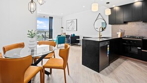 A modern kitchen with black cabinets, a central island, and a dining area with a glass table and orange chairs. - Stay Luxe Southport by Coastal Letting Company (Southport)
