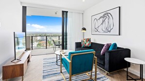A modern living room with a black sofa, a teal armchair, and a small round table. A large window offers a city view. - Stay Luxe Southport by Coastal Letting Company (Southport)