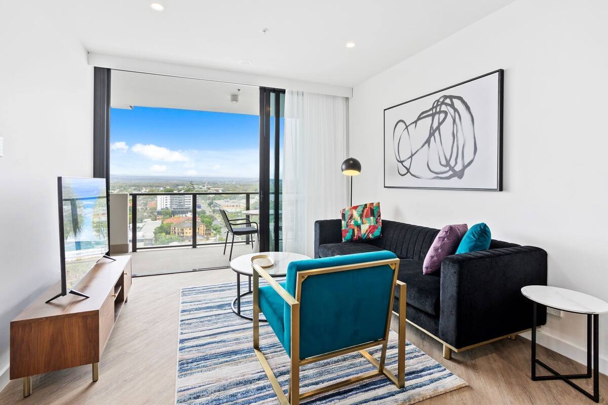 A modern living room with a black sofa, a teal armchair, and a small round table. A large window offers a city view.