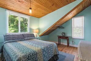 Ein Schlafzimmer mit geneigter Decke, einem Bett mit gemustertem Überwurf, einem Nachttisch mit Lampe und einer Pendelleuchte.