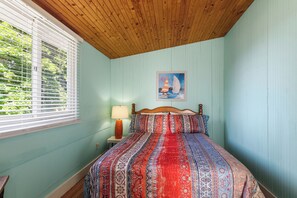 Een slaapkamer met een houten plafond, een bed met beddengoed met een patroon, een nachtkastje met een lamp, en een schilderij van een zeilboot aan de muur.
