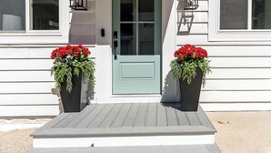 Uma casa com porta verde, dois vasos com flores vermelhas e um alpendre de madeira.