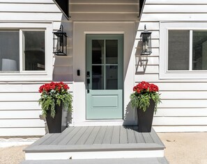 Ein Haus mit einer grünen Tür, zwei mit roten Blumen bepflanzten Blumentöpfen und einer Holzveranda.