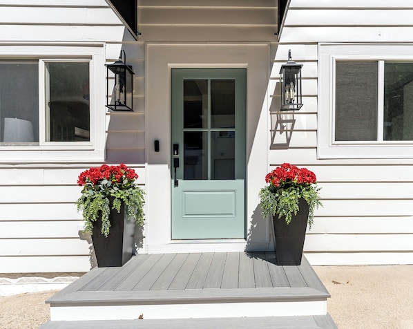Een huis met een groene deur, twee bloempotten met rode bloemen en een houten veranda.