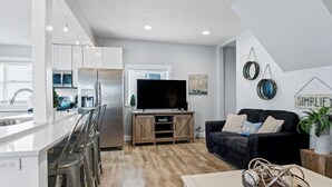 Eine moderne Küche mit großem Fernseher, ein Essbereich mit Barhocker und ein Wohnzimmer mit schwarzem Sofa und Couchtisch.