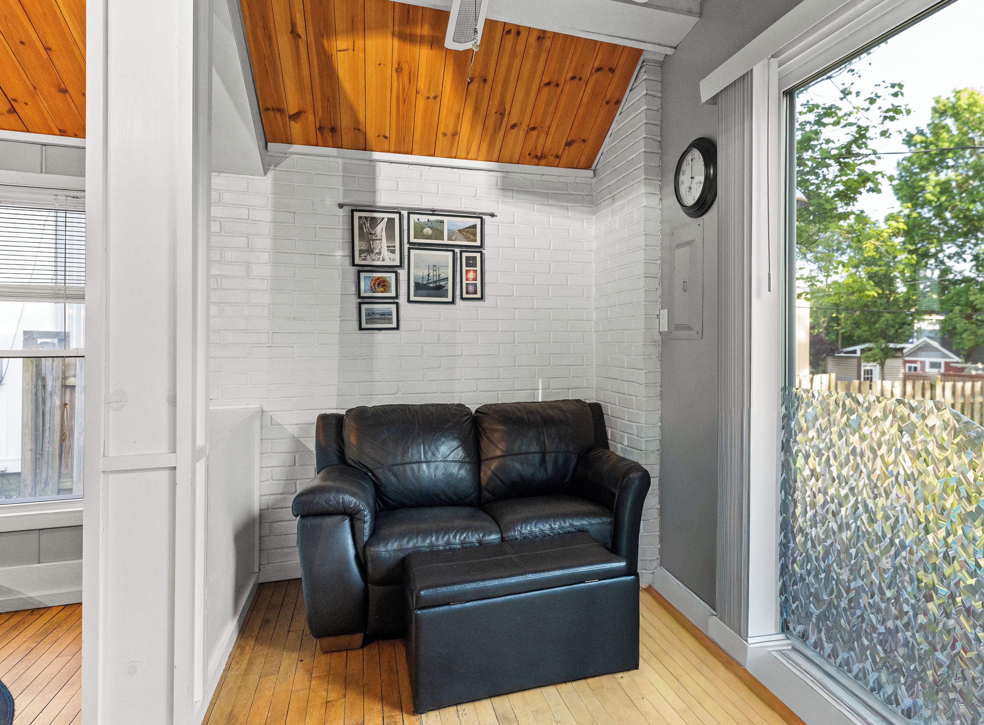 A room with a white brick wall, a black leather sofa, a wooden ceiling, and a large window with a view of trees.