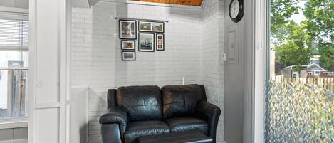 A room with a white brick wall, a black leather sofa, a wooden ceiling, and a large window with a view of trees.