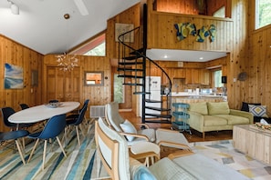 A wooden cabin interior with a dining area, a sofa, a staircase, and a kitchen.