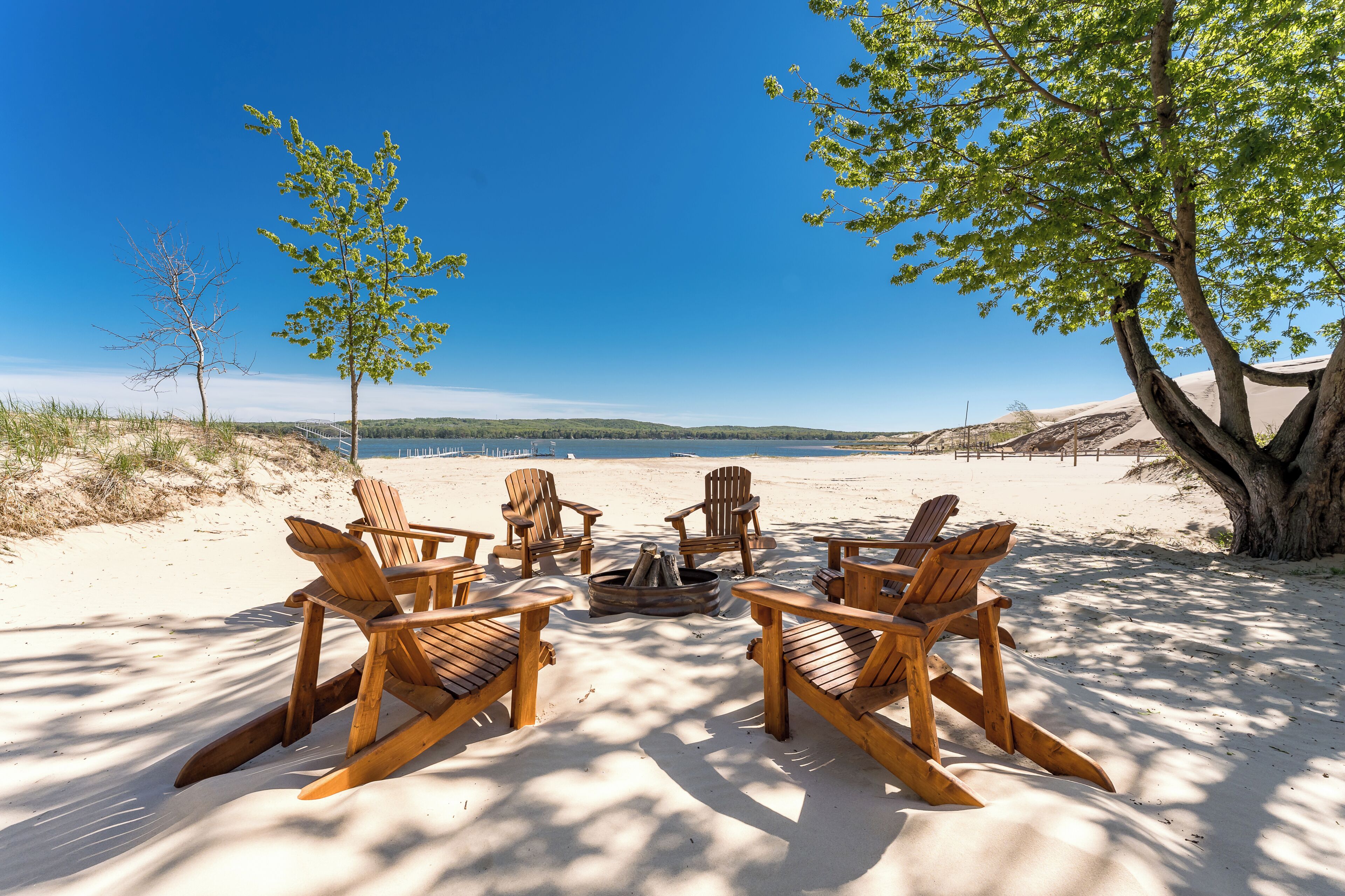 A beach setting with wooden chairs arranged around a fire pit, a tree, and a clear view of the water.