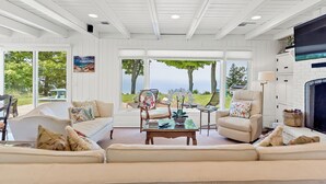 A spacious living room with a large sofa, two armchairs, a coffee table, and a fireplace. There are large windows offering a view of the outdoors.