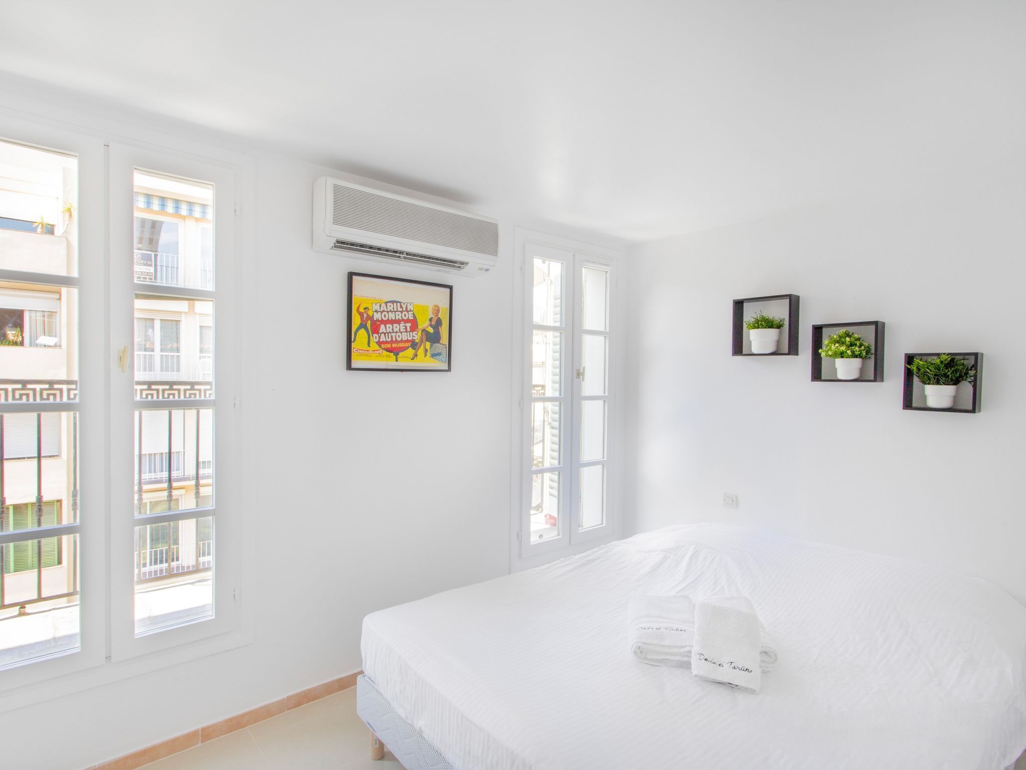 A bright, white room with a bed, a poster on the wall, and three potted plants.