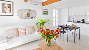 Una sala de estar moderna con un sofá blanco, una mesa de comedor con sillas, un jarrón de flores y una cocina con armarios blancos y una cafetera.