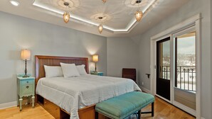 A bedroom with a large bed, wooden headboard, two bedside tables with lamps, a bench, and a sliding glass door to a balcony. - Downtown Grand Haven Stay | Spacious 2-Bed in Former Theatre (Grand Haven)