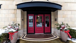 A hotel entrance with a red door, a black awning, and two large potted plants with red ribbons. - Downtown Grand Haven Stay | Spacious 2-Bed in Former Theatre (Grand Haven)