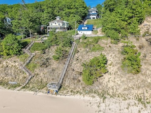 Una propiedad costera con una escalera que baja a una playa, rodeada de un bosque denso y una casa grande con techo azul.