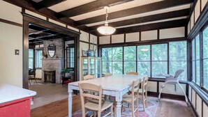 Un comedor con una mesa grande y sillas, chimenea y ventanas amplias con vista a árboles.