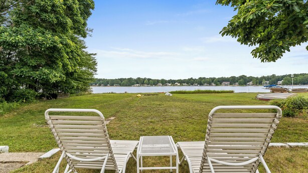 Two white metal chairs and a small table on a grassy lawn with a view of a lake and boats in the background. - Charming 2-Bedroom Strong Cottage in Twin Lake, MI (Twin Lake)