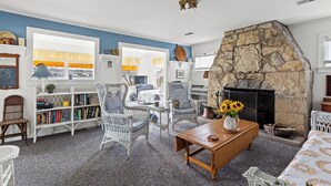 A cozy living room with a stone fireplace, wicker furniture, and a bookshelf filled with books. - Charming 2-Bedroom Strong Cottage in Twin Lake, MI (Twin Lake)