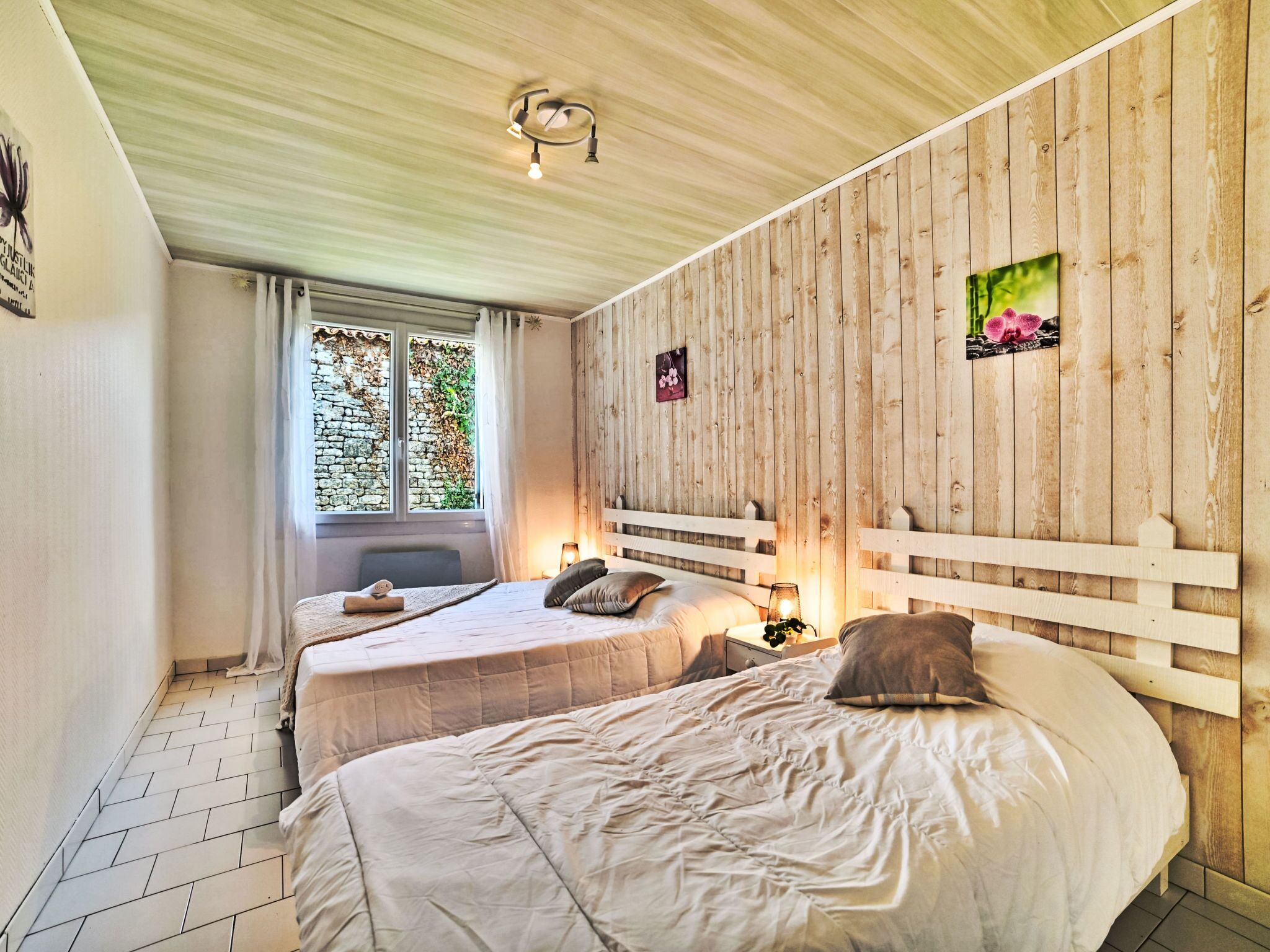 A bedroom with two beds, wooden panel walls, a stone wall visible through a window, and a small picture on the wall.